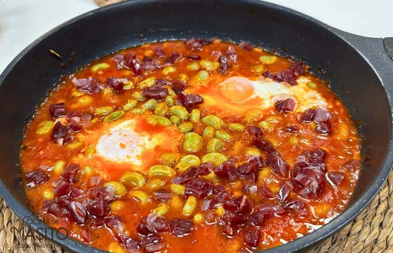 Plato de habas con jamón y huevo frito, servido en una sartén rústica, ideal para una comida rápida y deliciosa.