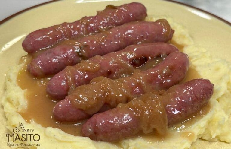 Salchichas doradas en salsa de cerveza acompañadas de cebolla caramelizada en un sartén.