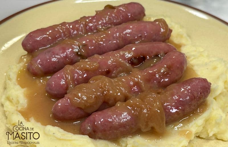 Salchichas doradas en salsa de cerveza acompañadas de cebolla caramelizada en un sartén.